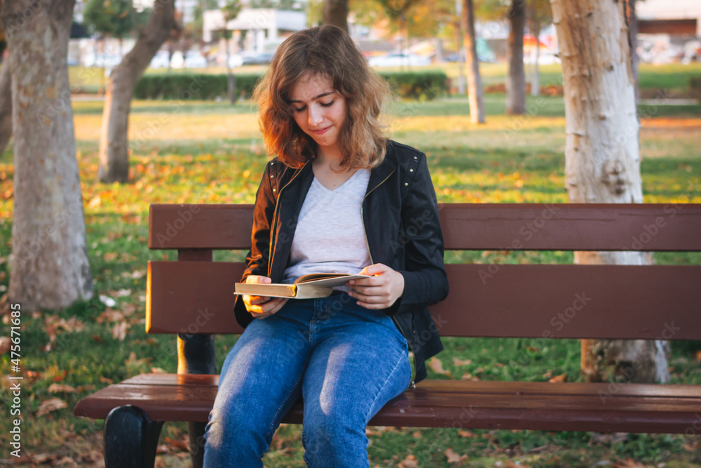Obraz premium Cheerful teenager girl reading book on the bench at autumn park
