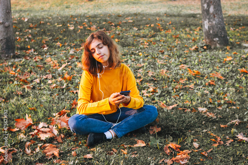 Young girl in yellow sweater listening to the music with earphones sitting on autumn grass