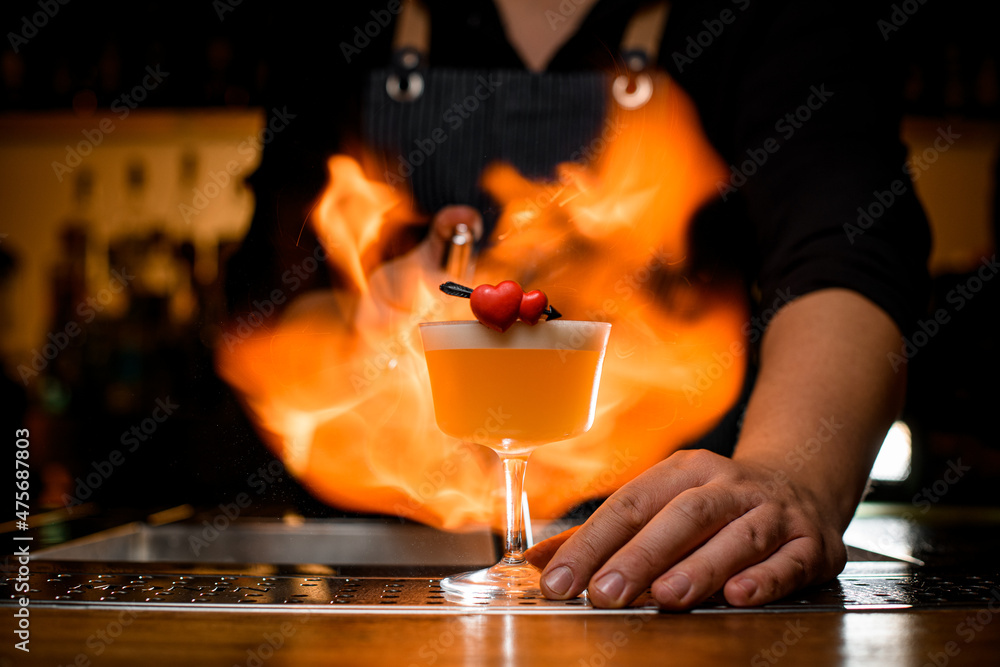 Bartender impressively sets fire on glass with whiskey sour cocktail decorated with two small red hearts Bartender impressively sets fire on glass with whiskey sour cocktail decorated with two small red hearts