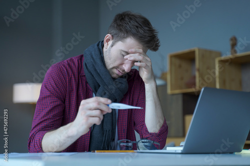 Unhappy austrian man freelancer checking body temperature at work, holding thermometer