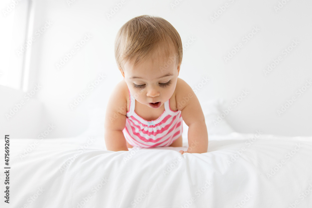 Cute baby girl crawling on white bed looking down with surprised expression Cute baby girl crawling on white bed looking down with surprised expression