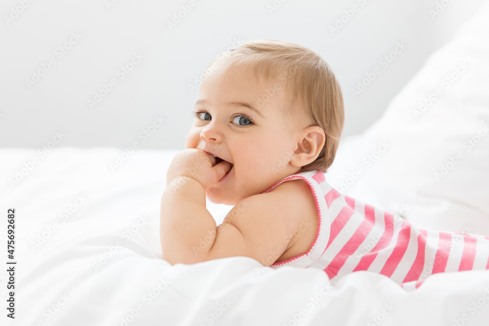Smiling baby girl lying on white bed with hand in mouth Smiling baby girl lying on white bed with hand in mouth