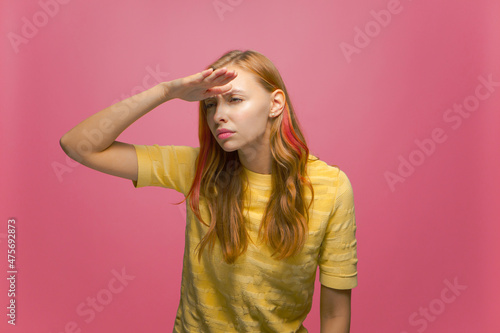 Focused attentive serious young woman looking far away with hand over head, searching on pink studio background