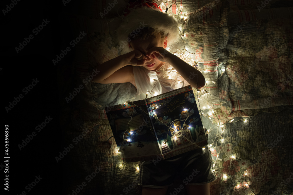 A sleepy kid with a Christmas book under small lights dreaming with a Merry Christmas full of gifts and joy.