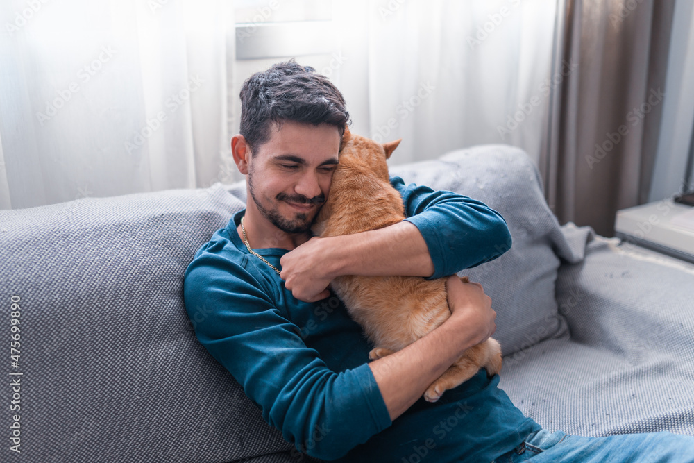 young man sitting on a gray sofa lovingly hugs a brown tabby cat