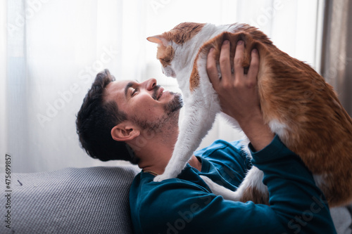young man sitting on a gray sofa holds in his arms a brown and white cat 