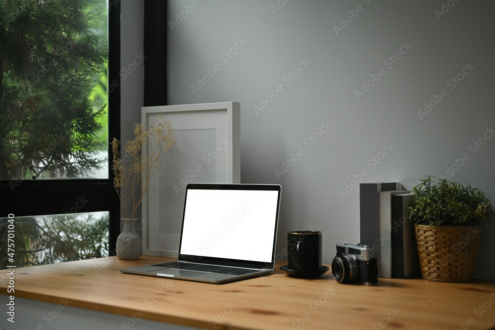 Photo of a comfortable workplace surrounded by a white blank screen computer laptop, empty picture frame, wicker basket, vintage camera, coffee cup, jar and books. Photo of a comfortable workplace surrounded by a white blank screen computer laptop, empty picture frame, wicker basket, vintage camera, coffee cup, jar and books.