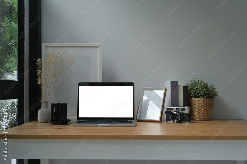 Photo of a comfortable workplace surrounded by a white blank screen computer laptop, empty picture frame, wicker basket, vintage camera, coffee cup, jar and books. Photo of a comfortable workplace surrounded by a white blank screen computer laptop, empty picture frame, wicker basket, vintage camera, coffee cup, jar and books.