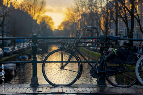 Selective focus front wheel of classic bicycle and handlebar on canal in the evening during sunset, Blurred traditional houses, Amsterdam, Netherlands, Cycling is a common mode of transport in Holland #475701819