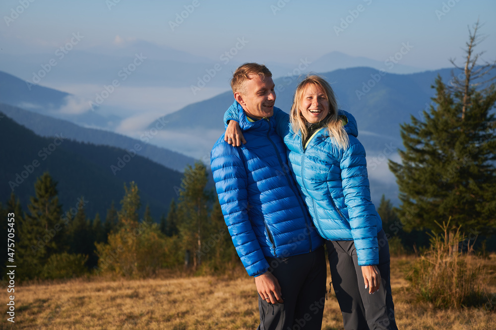 Two happy travelers standing and embracing on the background of trees, mountain hills, covered by forests and silhouettes mountain peaks far away. Nice active hiking in the mountains.