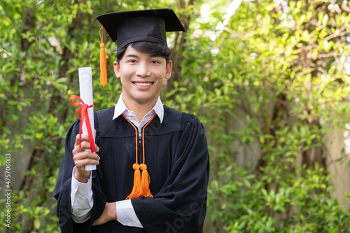 In the graden, a happy young Asian man wears graduation gowns and has a diploma.
