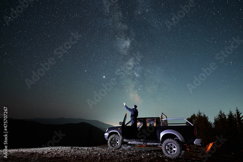 Person standing in the car and looking at stars at night. His left arm outstretched as if in welcome with sky and Milky Way galaxy. Starry beauty over mountain beskids on the background.