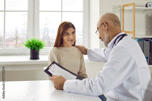 Friendly, supportive senior male doctor talking to his female patient. Adult physician sitting at his working desk and reassuring happy young woman who came to hospital for medical consultation