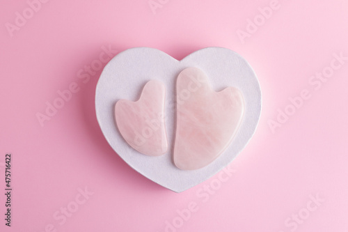 Rose quartz gua sha stones for face and body relaxation and lifting massage. Acupressure massagers on decorative paper heart isolated on pastel pink background.