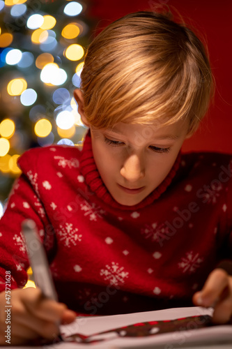 a boy in a red Christmas sweater on the background of a Christmas tree bent over the drawing in the process of decorating it with a marker