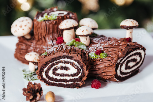 French dessert called Yule log or bûche de Noël with merengue mushrooms and mint leaves on top of chocolate glazing. Placed in front of Christmas tree. Decorated for Christmas Holidays or New year