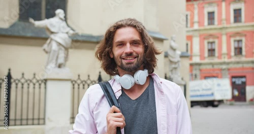Good-looking male student with headphones holding bag and standing on street in city. Portrait of young Caucasian guy turning head, looking to camera and sincerely smiling. Technology concept.