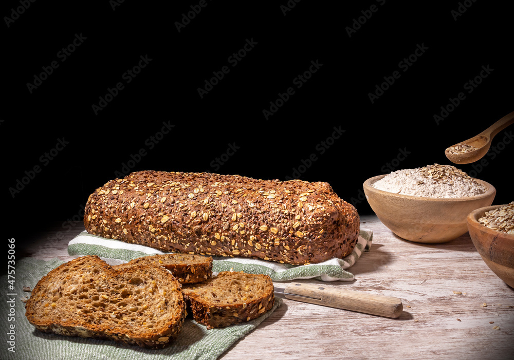 Still life of protein bread. Whole and sliced ​​protein bread, on black background on wooden table, with wooden bowl full of flour and wooden spoon with multiple cereals