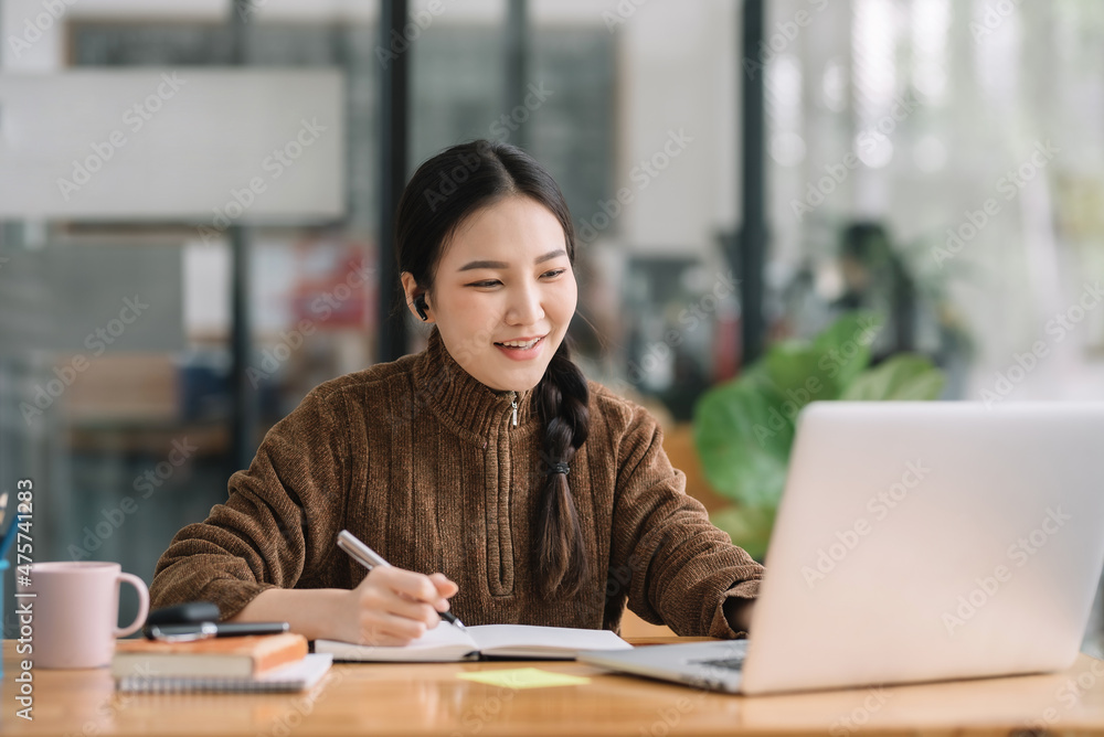 Young Asian female student sitting taking notes and using laptop computer at table. Young Asian female student sitting taking notes and using laptop computer at table.