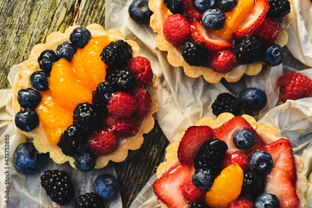 Above tabletop view of four inch tarts with custard filled baked pastry crust shells topped with glazed blueberries, red raspberries, blackberries, sliced strawberries, orange sections Above tabletop view of four inch tarts with custard filled baked pastry crust shells topped with glazed blueberries, red raspberries, blackberries, sliced strawberries, orange sections