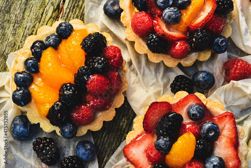 Above tabletop view of four inch tarts with custard filled baked pastry crust shells topped with glazed blueberries, red raspberries, blackberries, sliced strawberries, orange sections