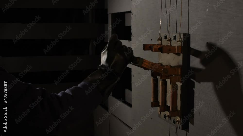 In dramatic lighting a black rubber gloved hand reaches into the frame to grab the wooden handle of an open old large industrial electrical bar switch then hesitates and prepares before shoving it on.
