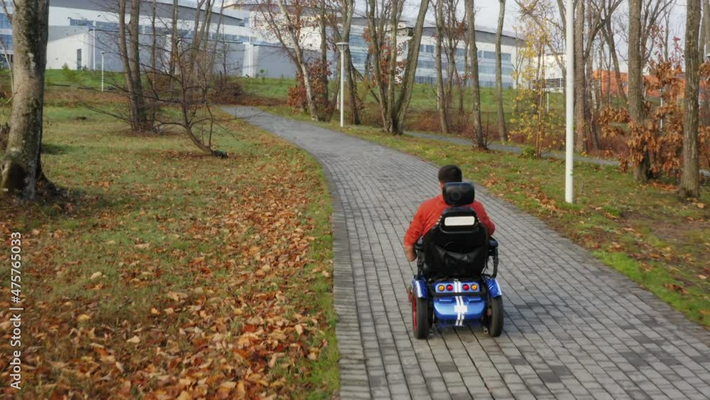 Aerial zooming in view of a caucasian man living with a disabiity with amputated legs and arms driving on electric wheelchair along the empty path in autumn park Aerial zooming in view of a caucasian man living with a disabiity with amputated legs and arms driving on electric wheelchair along the empty path in autumn park