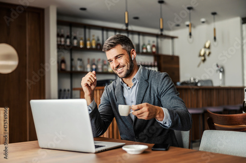 A happy businessman is sitting in a cafe, drinking coffee and watching a webinar on the laptop. The man has earphones in his ears so he can listen to the lecture. A businessman using laptop in cafe © dusanpetkovic1