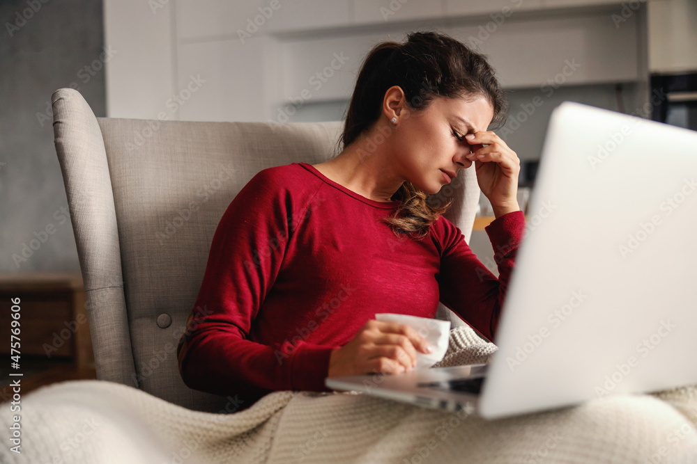 Ill woman sitting in chair at home during lockdown with laptop in her lap and having headache. Ill woman sitting in chair at home during lockdown with laptop in her lap and having headache.