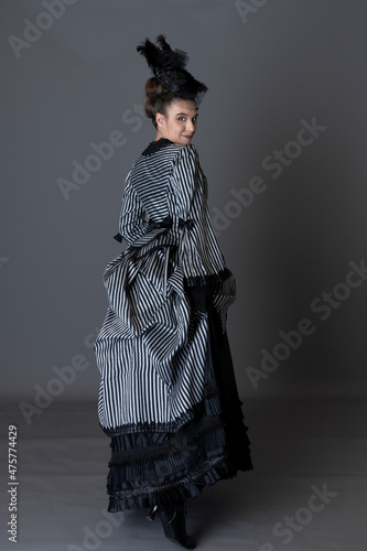 A Victorian woman wearing a striped silk polonaise and black skirt alone against a studio backdrop