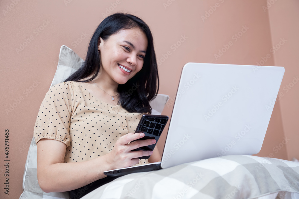 Beautiful woman on bed working with laptop while holding cell phone Beautiful woman on bed working with laptop while holding cell phone