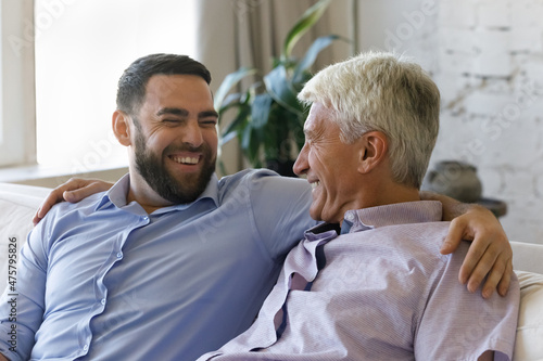 Happy laughing young handsome man embracing shoulders of sincere joyful middle aged senior retired father, having fun joking or involved in trustful conversation sitting on cozy couch at home.