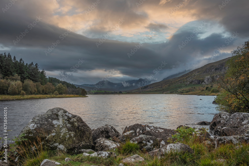 Epic stunning Autumn landscape image of Snowdon Massif viewed from shores of Llynnau Mymbyr at sunset with dramatic dark sky and clouds