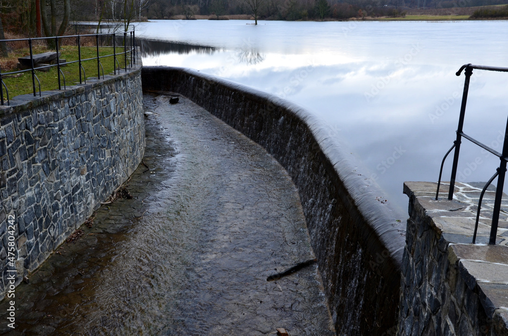 bridge over safety spillway of the dam. stone bridge with natural paving of gray flat stones. on the edge of the retaining walls is a subtle wrought iron railing of color. water flows under through