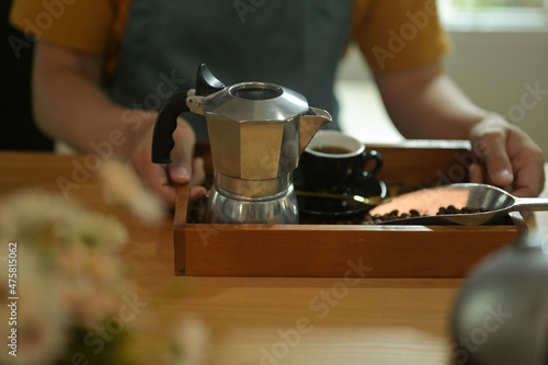 Photo of waiter's hand holding a serving tray full of coffee bean, coffee cup, steel kettle and coffee bean scoop. © saltdium