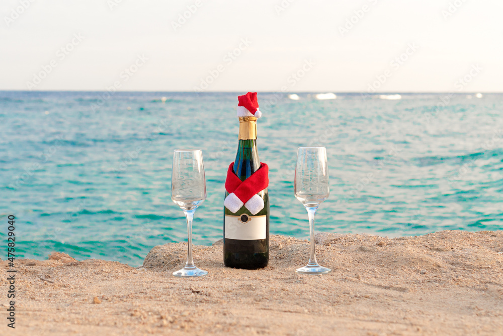 A cheerful bottle of champagne in a Santa Claus hat and two glasses on the beach against the background of the sea. Christmas concept. A cheerful bottle of champagne in a Santa Claus hat and two glasses on the beach against the background of the sea. Christmas concept.