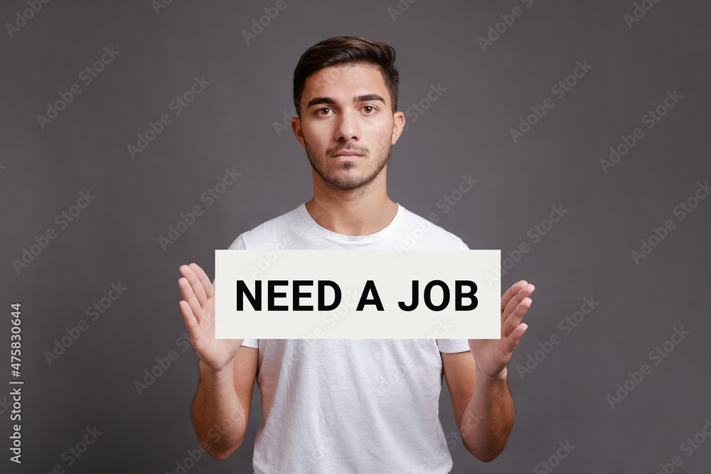 Handsome caucasian man holding sheet of paper with text need a job while standing isolated over gray background, looking for a work, job search concept, toned image Handsome caucasian man holding sheet of paper with text need a job while standing isolated over gray background, looking for a work, job search concept, toned image