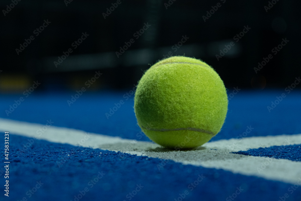 A paddle tennis ball in the foreground on the line of a blue paddle tennis court at night.