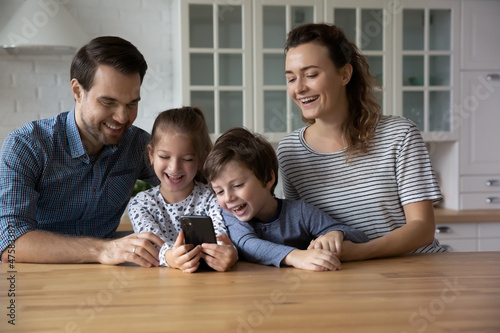 Happy parents with two kids using smartphone at home together, family having fun with mobile device, smiling Caucasian mother and father with little son and daughter looking at phone screen
