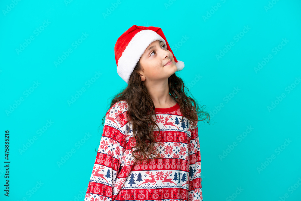 Little caucasian with christmas hat isolated on blue background looking up while smiling Little caucasian with christmas hat isolated on blue background looking up while smiling