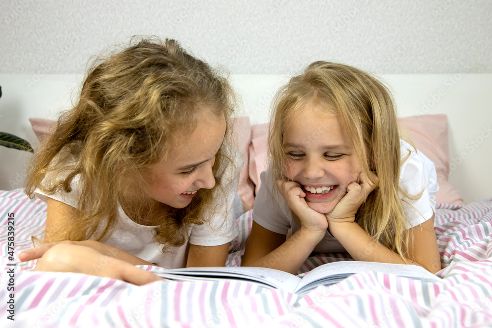 two cute sisters are lying on the bed and reading a funny book, smiling and laughing, family evening, one looking at the other, children's development through independent reading two cute sisters are lying on the bed and reading a funny book, smiling and laughing, family evening, one looking at the other, children's development through independent reading