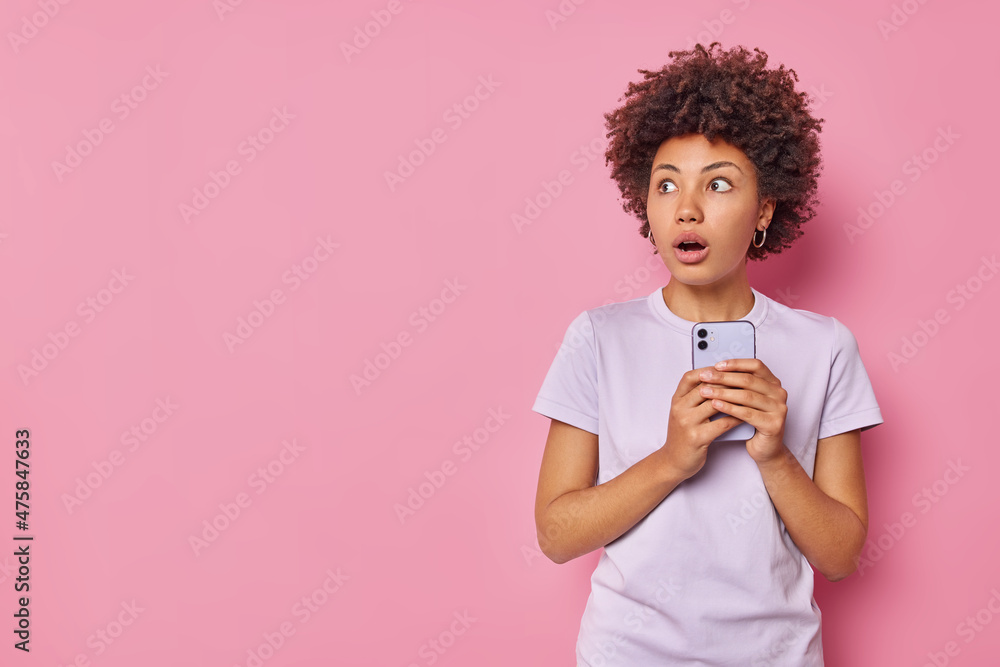 Horizontal shot of scared stunned woman holds mobile phone sees something scary wears casual t shirt isolated over pink background with copy space for your advertising content hears bad news