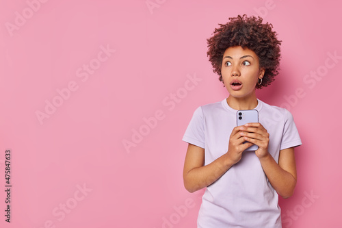 Horizontal shot of scared stunned woman holds mobile phone sees something scary wears casual t shirt isolated over pink background with copy space for your advertising content hears bad news