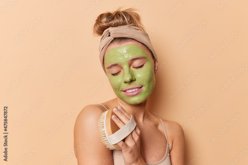 Charming woman exfoliates skin pampers herself makes massage with dry brush applies green moisturising mask wears headband stands bare shoulders isolated over beige studio wall. Body scrubbing