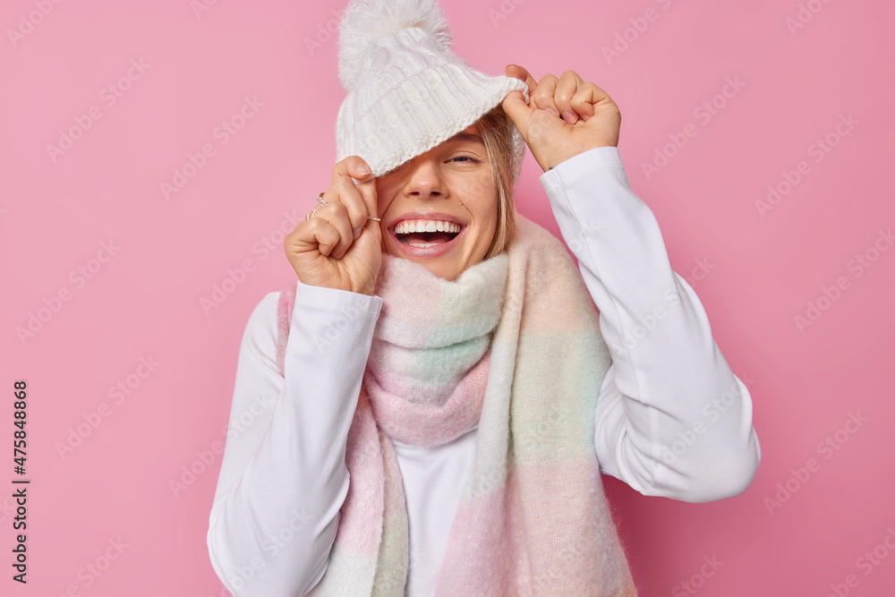Joyful teenage girl has fun hides face behind white knitted hat has upbeat mood wears sweater and scarf around neck smiles broadly has perfect even teeth foolishes around being full of energy Joyful teenage girl has fun hides face behind white knitted hat has upbeat mood wears sweater and scarf around neck smiles broadly has perfect even teeth foolishes around being full of energy