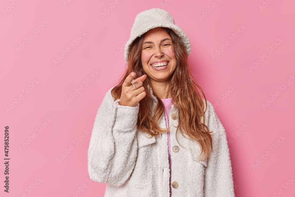 Joyful young woman long long wavy hair points index finger at you notices something funny in front smiles gladfully wears fur coat and panama isolated over pink studio background. Hey join me Joyful young woman long long wavy hair points index finger at you notices something funny in front smiles gladfully wears fur coat and panama isolated over pink studio background. Hey join me