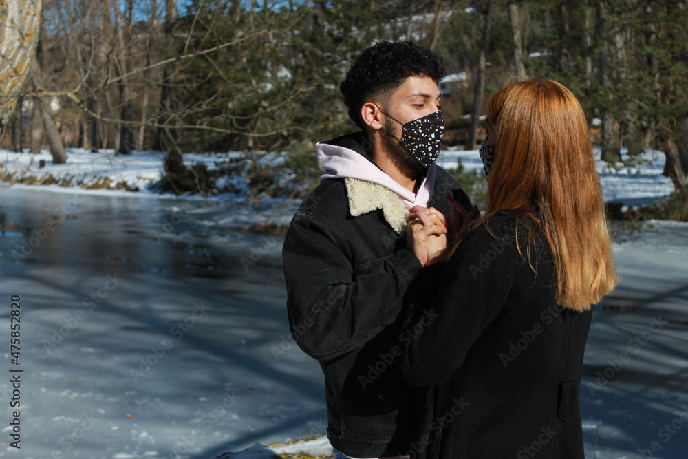 young interracial couple in masks hugging and looking at each other. romantic journey through the snow by an icy river. young interracial couple in masks hugging and looking at each other. romantic journey through the snow by an icy river.