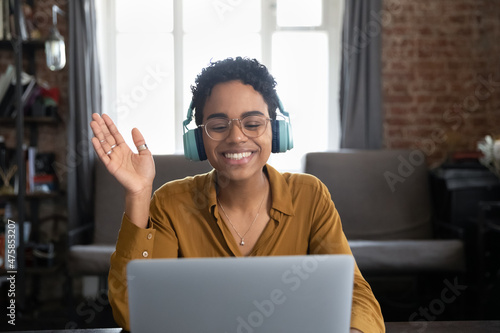 Happy attractive millennial African American woman in eyewear and headphones waving hand looking at laptop screen, starting distant video conference call, communicating remotely at home office. photo