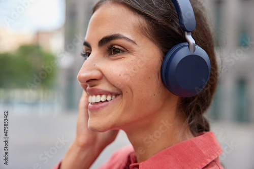 Sideways shot of happy brunette young woman listens audio track via headphones smiles broadly stands in profile against blurred background strolls outdoors. People hobby and lifestyle concept