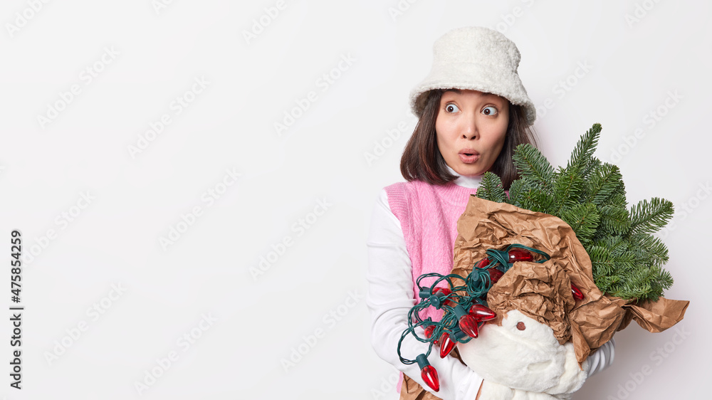 Obraz premium Horizontal shot of shocked brunette woman wears white winter panama and warm mittens looks amazed away holds New Year attributes for decoration isolated over white background with blank copy space